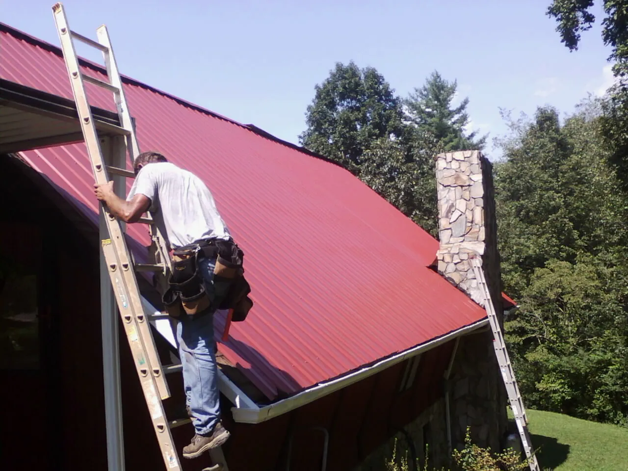 Roofer on a ladder beside a red metal roof and stone chimney — Soffit & Fascia Repair by Roofing Brothers in Baton Rouge
